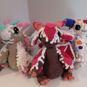 May include: Three crocheted stuffed dragons in various colors, including pink, red, white, and blue. The dragons have large eyes and are sitting on a white surface.