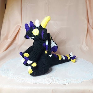May include: A crocheted dragon plush toy with black, yellow, white, and purple yarn. The dragon is sitting on a white lace doily.