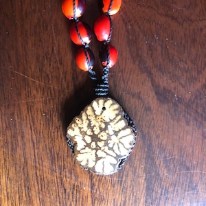 Può includere: Una collana fatta a mano con un ciondolo con un motivo floreale e una fila di perline rosse e nere. Il ciondolo è marrone con un motivo color crema e le perline sono rosse e nere. La collana è infilata su un cordino nero.