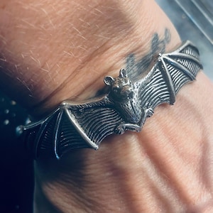 May include: A silver-toned metal cuff bracelet featuring a detailed, sculpted bat with outstretched wings. The bat is positioned in the center of the band, with its wings extending along the sides of the wrist. The metal has a dark, oxidized finish.