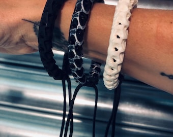 Peut inclure: Trois bracelets réglables en noir, noir et blanc et blanc cassé. Le bracelet noir a une surface texturée. Le bracelet noir et blanc a un motif tacheté. Le bracelet blanc cassé a un design en forme d'os. Chaque bracelet a des cordons noirs.