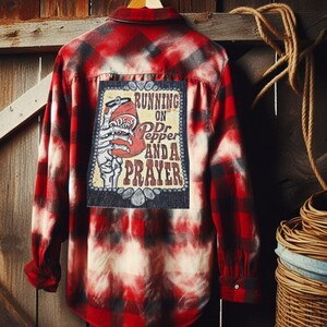 May include: Red and black plaid flannel shirt with a bleached design. The back features a patch with a Dr. Pepper can held by a skeleton hand, and the text "RUNNING ON Dr Pepper AND A PRAYER". The shirt is hanging on a wooden hanger.