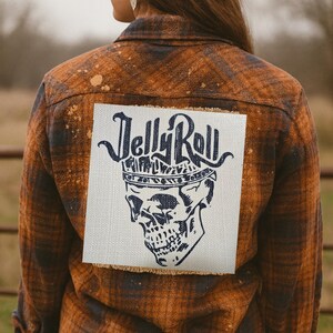 Peut inclure: Chemise en flanelle à carreaux marron et marine avec un design vieilli. Le dos de la chemise présente un écusson carré avec les mots "Jelly Roll" au-dessus d'un crâne portant une couronne. La chemise est portée avec une casquette de baseball gris foncé.