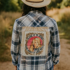 May include: A blue and white plaid flannel shirt with a patch featuring a portrait and the text "Tease it to Jesus and spray it like hell." The shirt is worn with a straw cowboy hat.