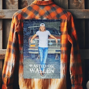 May include: Orange and black plaid flannel shirt with a graphic on the back. The graphic features a person in a white t-shirt and blue jeans, leaning against a red truck. The text "WASTED ON WALLEN" is below the image.