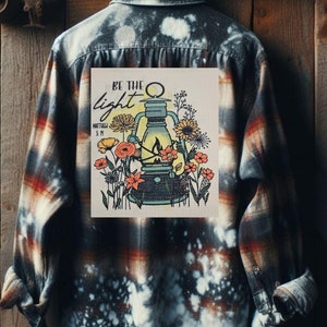 May include: A long-sleeved flannel shirt with a bleached design. The back of the shirt features a patch with a lantern and floral design, with the words "BE THE light" and "Matthew 5:19". The shirt is hanging on a wooden background.