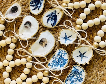 May include: A collection of oyster shell ornaments with various blue and white designs, including a starfish, floral patterns, and a sea turtle. The ornaments are strung on a wooden bead garland with gold accents.