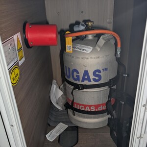 Op de afbeelding: Een zilveren propaangasfles, met het label "UGAS", is vastgezet in een compartiment. De fles heeft zwarte banden en een rode slang. Een rode cilinder en waarschuwingslabels zijn links zichtbaar. Het compartiment is van hout.