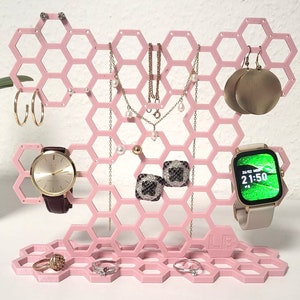 May include: Pink honeycomb-shaped jewelry organizer with a watch stand, a watch, necklaces, earrings, and rings. The organizer has a base with hexagonal compartments.