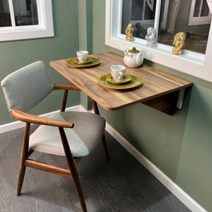 May include: A light wood folding wall table with a white chair. The table has two place settings with green and white plates and cups. There are three small gold and silver figurines on the windowsill behind the table.