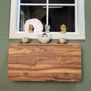 May include: A wooden folding table with a light brown wood grain finish. The table is mounted to a wall and has a white window above it. There are three small decorative figurines and two teacups on the table.
