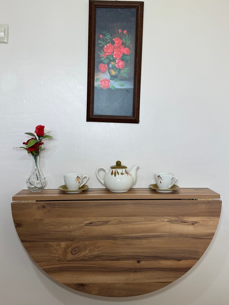 Pu&ograve; includere: Un tavolo pieghevole a mezza luna in legno con una teiera, due tazze da t&egrave; e un vaso di fiori. Un'immagine incorniciata di rose &egrave; appesa sopra il tavolo. Il tavolo &egrave; in legno con venature naturali.