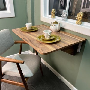 May include: A small wooden table with a white chair, a teapot, two cups, and two saucers. The table is mounted to the wall and has a light wood finish. The chair is upholstered in a light gray fabric.