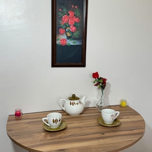 Pu&ograve; includere: Una natura morta con un quadro incorniciato di rose rosse in un vaso, una teiera bianca con coperchio verde, due tazze e piattini, un piccolo vaso con fiori rossi e due candele. Su un tavolo di legno a semicerchio.
