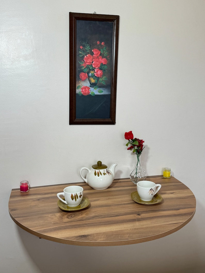 Pu&ograve; includere: Una natura morta con un quadro incorniciato di rose rosse in un vaso, una teiera bianca con coperchio verde, due tazze e piattini, un piccolo vaso con fiori rossi e due candele. Su un tavolo di legno a semicerchio.