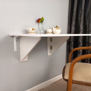 Pu&ograve; includere: Tavolo pieghevole bianco a parete con un piano bianco e due staffe metalliche bianche. Il tavolo ha una teiera, due tazze e un vaso con fiori rossi. Una sedia in legno con un cuscino beige &egrave; in primo piano.