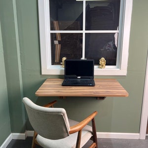 May include: A wooden desk with a laptop computer and a chair. The desk is mounted to the wall and has a light wood finish. The chair is upholstered in a light gray fabric and has a wooden frame.