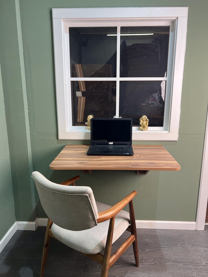 May include: A wooden desk with a laptop computer and a chair. The desk is mounted to the wall and has a light wood finish. The chair is upholstered in a light gray fabric and has a wooden frame.