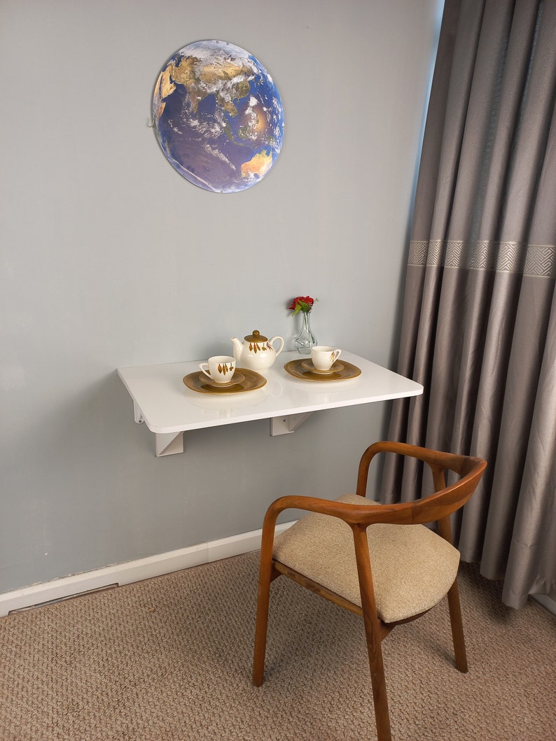 Pu&ograve; includere: Un tavolo pieghevole bianco con un globo sul muro dietro di esso. Ci sono una teiera e due tazze sul tavolo, insieme a un vaso di fiori rossi. Una sedia di legno con un cuscino beige si trova accanto al tavolo.