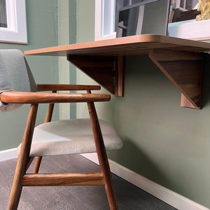 May include: A light wood folding desk with a laptop on top, mounted to a green wall. A light wood chair with a light gray cushion sits in front of the desk.
