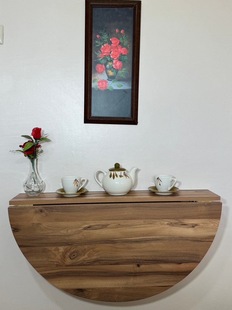Pu&ograve; includere: Un tavolo a ribalta in legno, montato a parete, con un design a semicerchio. Una teiera bianca con coperchio verde e due tazze da t&egrave; con piattini sono sul tavolo. Sono visibili anche un dipinto floreale incorniciato e un vaso con una rosa rossa.