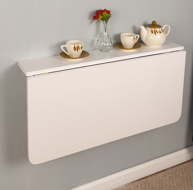 Pu&ograve; includere: Tavolo pieghevole a parete bianco con un piano bianco e una base bianca. Il tavolo &egrave; ripiegato e ha un piccolo vaso con fiori rossi in cima. Ci sono due tazze da t&egrave; bianche e una teiera bianca sul tavolo.