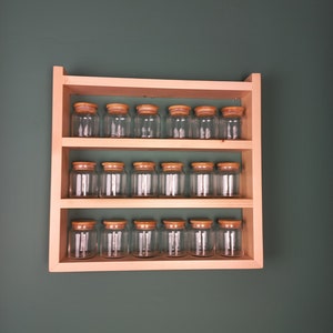 Puede incluir: Un estante de especias de madera con tres estantes, cada uno con seis tarros de vidrio transparentes con tapas de madera. El estante está montado en una pared verde.