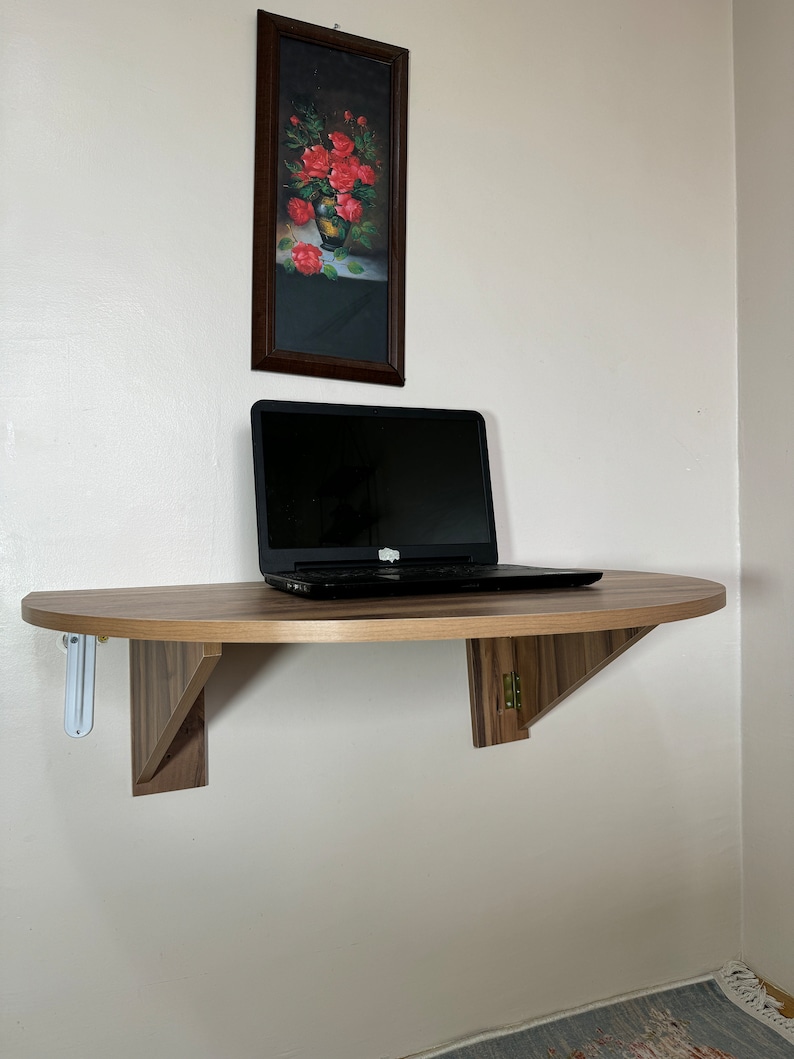 Pu&ograve; includere: Una scrivania in legno semicircolare montata a parete con un laptop sopra. La scrivania &egrave; sostenuta da due staffe triangolari. Sopra la scrivania c'&egrave; un dipinto incorniciato di rose rosse in un vaso. Il muro &egrave; di colore beige chiaro.