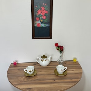 Pu&ograve; includere: Una natura morta con un tavolo semicircolare in legno con una teiera, due tazze e piattini e un vaso con una rosa rossa. Un dipinto incorniciato di rose &egrave; appeso sopra. Due piccole candele sono sul tavolo.