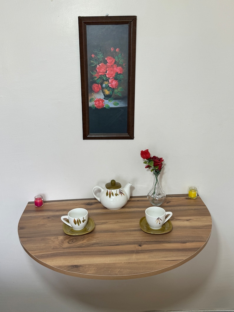 Pu&ograve; includere: Una natura morta con un tavolo semicircolare in legno con una teiera, due tazze e piattini e un vaso con una rosa rossa. Un dipinto incorniciato di rose &egrave; appeso sopra. Due piccole candele sono sul tavolo.