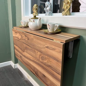 May include: A wooden folding wall table with a light brown wood grain finish. The table is folded up and mounted to a wall. There are three teacups and a teapot on the table.