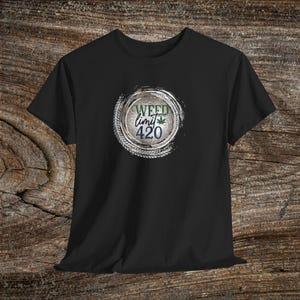 Puede incluir: Camiseta negra con un diseño circular plateado y verde con el texto "WEED limit 420" y una hoja de cannabis. La camiseta se muestra sobre una superficie de madera.