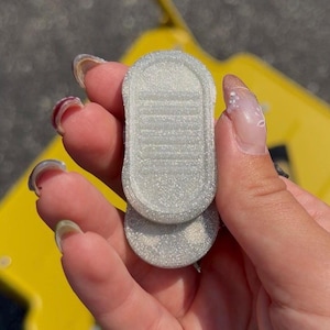 May include: A small, oval-shaped object with a glittery, silver-toned finish. The item features a raised, textured surface with horizontal lines and a smooth, rounded border. It has a compact, flat design with a slightly layered appearance.