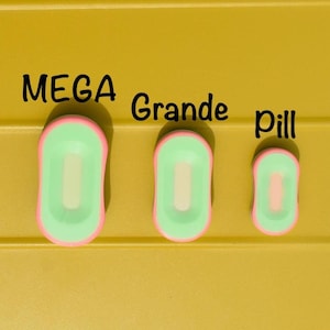 May include: Three pill-shaped objects in varying sizes, each with a light green interior and a pink border, are displayed against a yellow background. The text "MEGA Grande pill" is written above the objects.