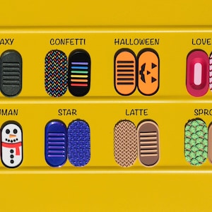 May include: Eight pairs of oval-shaped silicone slider covers arranged in two rows. Designs include Galaxy, Confetti, Halloween pumpkin, Lovebug hearts, Snowman, Star, Latte, and Sprout leaf patterns, each with a corresponding solid or textured base.