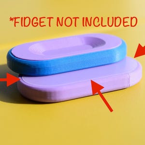 May include: Two stacked oval-shaped objects with smooth surfaces. The top object is blue with a lavender center insert featuring concentric ridges. The bottom object is solid lavender. Red arrows point to the objects, and text reads *FIDGET NOT INCLUDED.