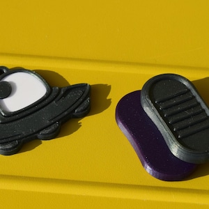 May include: Black and white UFO-shaped charm with a loop, beside a black and purple oval. The objects are set against a bright yellow background.