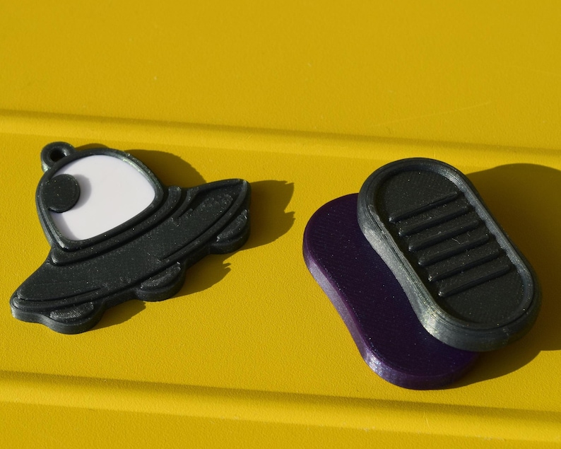 May include: Black and white UFO-shaped charm with a loop, beside a black and purple oval. The objects are set against a bright yellow background.