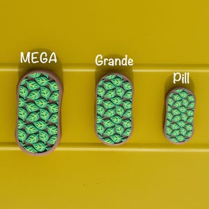 May include: Three leaf-patterned decorative items in varying sizes labeled "MEGA", "Grande", and "Pill". Each has a brown border and a green leaf design on a brown background. The items are displayed on a yellow surface.