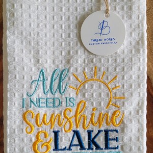 May include: White waffle weave kitchen towel with blue and yellow embroidery that reads "All I need is sunshine & lake water". A white circle tag with blue embroidery that reads "Thread Works Custom Embroidery" is attached to the towel.