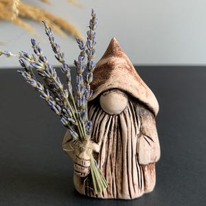 Puede incluir: Una figura de gnomo de cerámica con un gorro puntiagudo y una larga barba texturizada, sosteniendo un manojo de lavanda seca. El gnomo es de tonos marrones y beige, con una cara lisa y una pequeña bolsa. La lavanda tiene tonos morados y verdes.