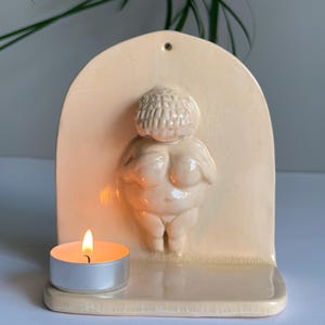 Puede incluir: Un portavelas de cerámica color crema con una figura de Venus esculpida. Una pequeña vela de té encendida se encuentra frente a la figura. El soporte tiene una parte superior arqueada con un pequeño agujero para colgar.