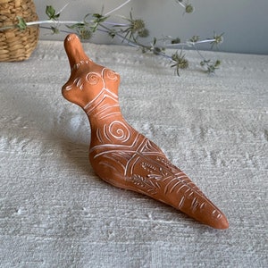 May include: A terracotta-colored figurine with white etched designs. The figure has a stylized form, with a rounded torso and elongated, pointed base. The white designs include spirals, lines, and leaf-like patterns. The figurine is likely a decorative art piece.