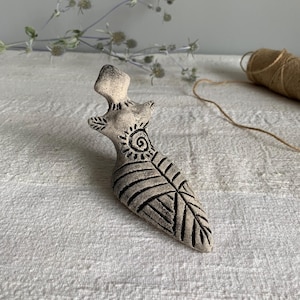 Puede incluir: Pequeña figura de cerámica blanquecina con forma de figura femenina estilizada. La figura presenta detalles grabados, incluyendo un diseño de sol y patrones de hojas. La pieza está sobre un fondo de tela blanca.