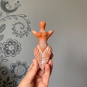 Puede incluir: Una figurita de terracota de una mujer con detalles pintados en blanco, incluyendo un símbolo solar en el pecho y un diseño de hoja en la falda.