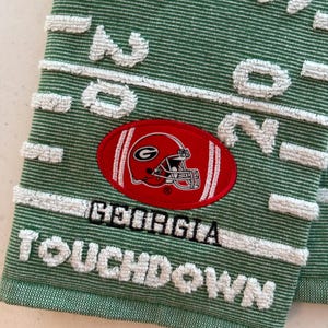 May include: A green textured towel featuring a white football field yard line pattern, an embroidered red oval patch with a Georgia Bulldogs helmet logo, and the embroidered text GEORGIA TOUCHDOWN in white and black lettering.