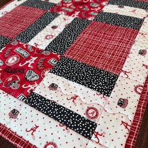 Könnte beinhalten: Eine handgefertigte Steppdecke mit einem Patchwork-Design in Rot, Schwarz und Weiß. Die Steppdecke enthält verschiedene Muster, darunter Punkte, Streifen und Stoff mit dem Wort "ALABAMA" und dem Buchstaben "A". Die Steppdecke hat einen roten Rand.