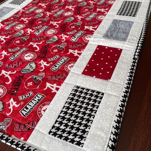 Könnte beinhalten: Ein roter, weißer und schwarzer Tischläufer mit einem Hahnentritt-Rand. Die Mitte des Läufers besteht aus rotem Stoff mit den Worten "Alabama" und "Bama", die in Weiß wiederholt werden. Der Läufer ist mit schwarzen und weißen Hahnentritt-Quadraten, weißen Tupfen-Quadraten und grauen Quadraten verziert.