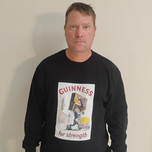 May include: Black long-sleeve shirt with a Guinness graphic. The graphic features the Guinness logo and the words "for strength" with an illustration of a man carrying a box. The shirt is a casual style.