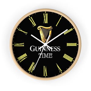 Op de afbeelding: Een zwarte en gouden wandklok met Romeinse cijfers en de tekst "GUINNESS TIME" op de wijzerplaat. De klok heeft een gouden harplogo met de tekst "ESTD 1759".